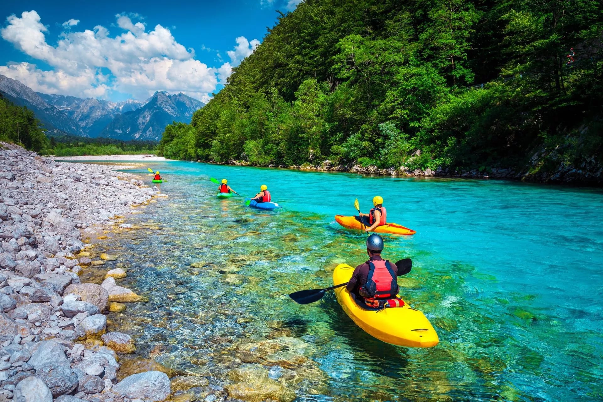 People kayaking on the clear turquoise Soca River with rocky banks and forested mountains.