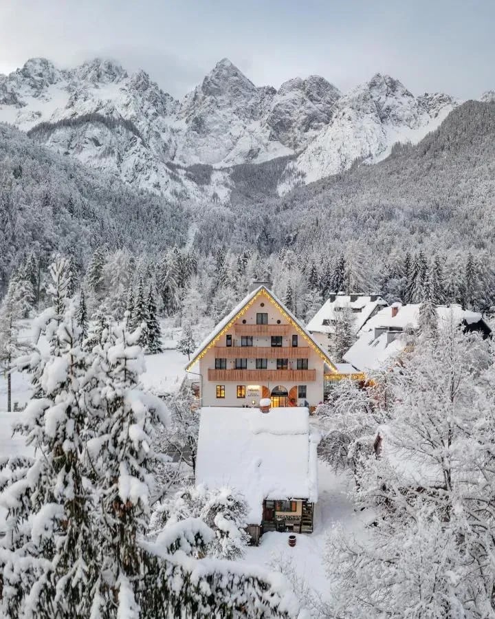 Alpine hotel with illuminated eaves surrounded by snow-covered trees and jagged, snowy mountains.