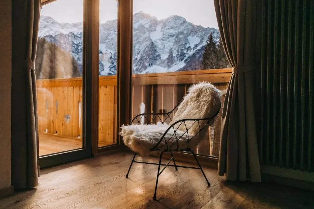 Chair with fur throw inside room overlooking snowy mountains from wooden balcony