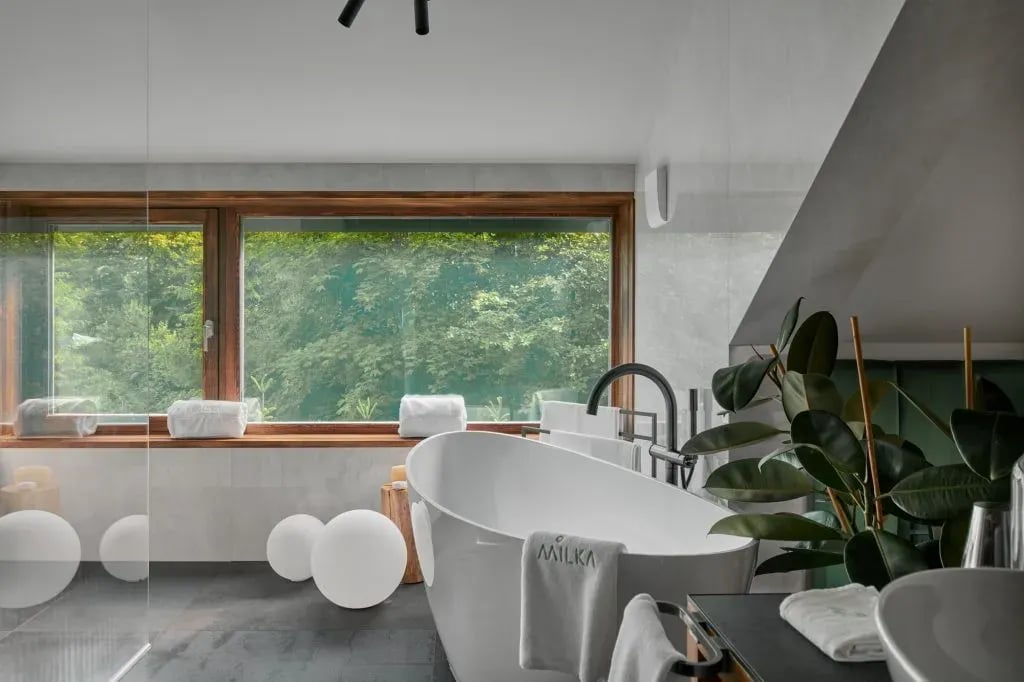 Modern bathroom with freestanding tub, large window overlooking lush green forest, and decorative orbs.