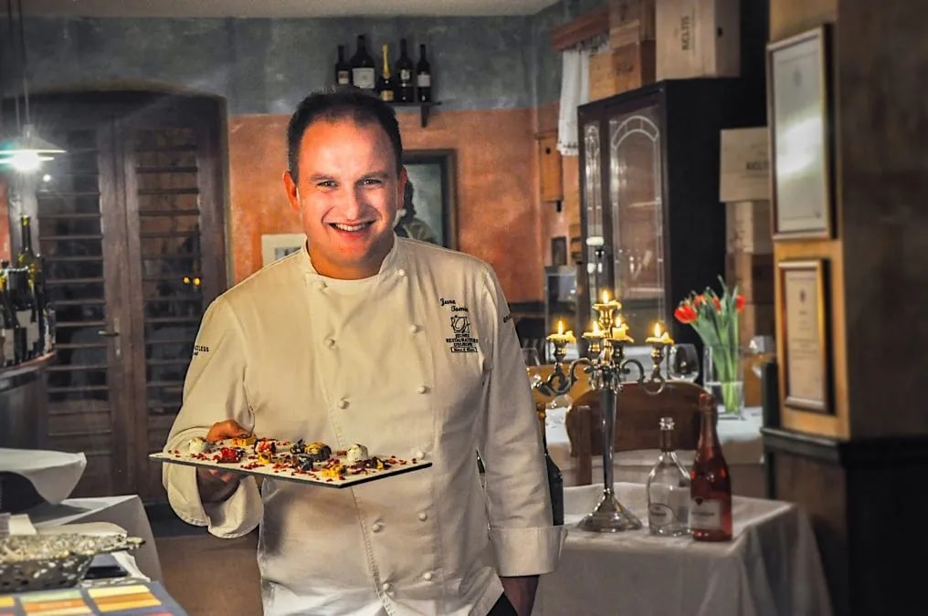 Chef Jure Tomic holding a plate of desserts in a warmly lit restaurant setting.