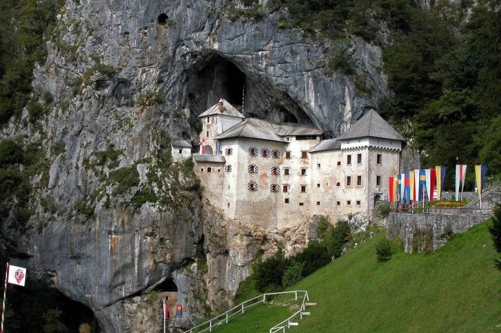 Predjama Castle built into a cliff face with colorful flags displayed on the green hillside.