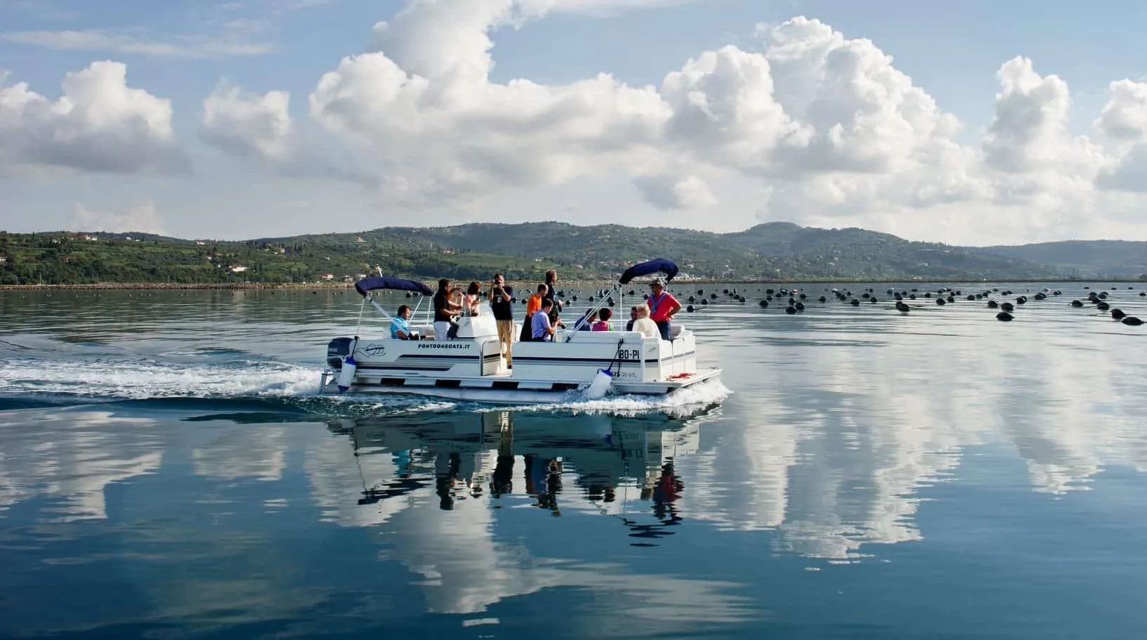 Pontoon boat carrying people moves across calm water near coastal hills and aquaculture buoys.