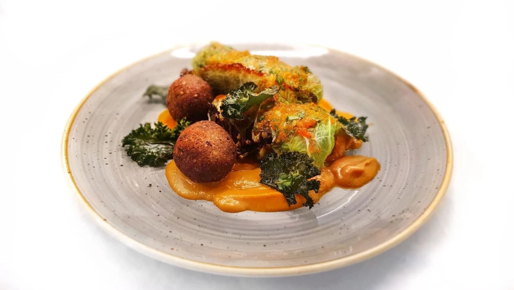 Cabbage rolls and fried balls served on orange puree with kale on a speckled plate