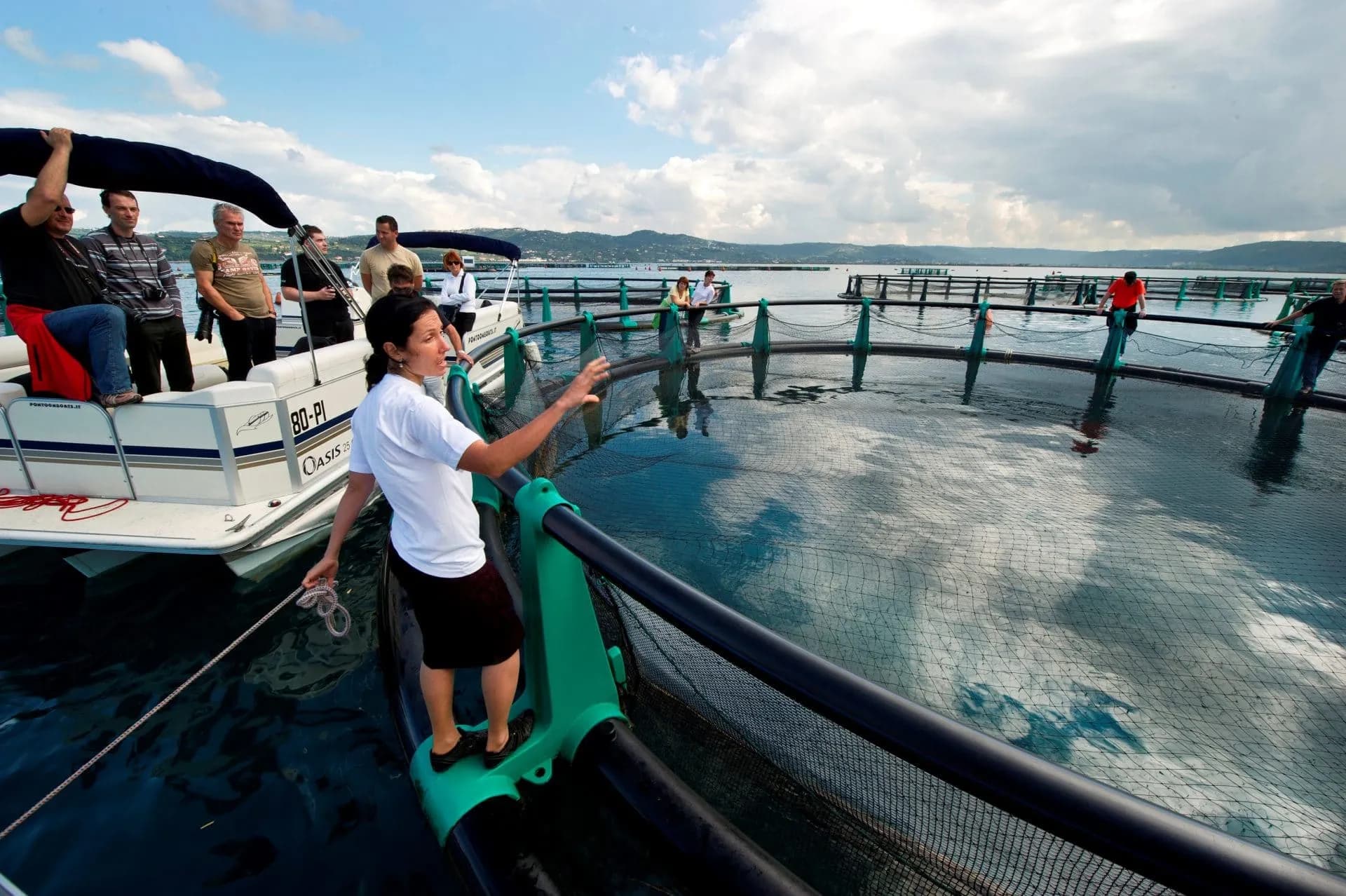Tour group on boat viewing circular fish farm enclosures on water with distant hills.