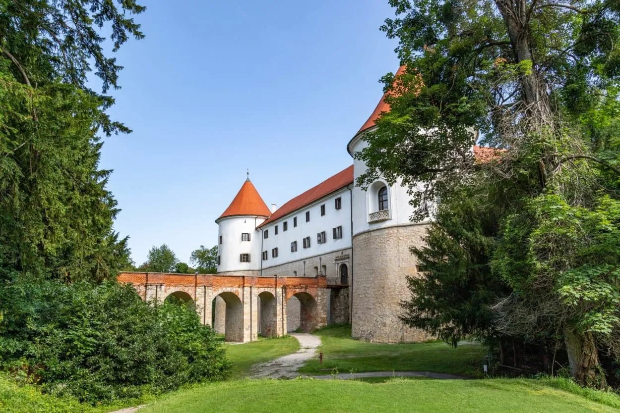 Brezice Castle with white walls, red roofs, and arched stone bridge surrounded by green trees.