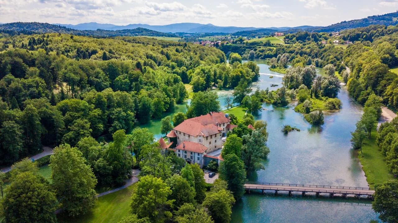 Aerial view of Hotel Grad Otočec on a river island surrounded by lush green forest and hills.