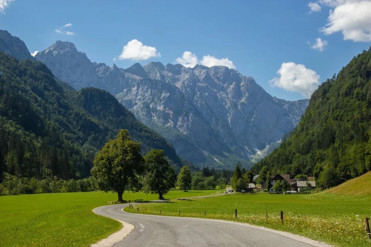 Winding road through green meadow valley towards Logar Valley mountains under blue sky.