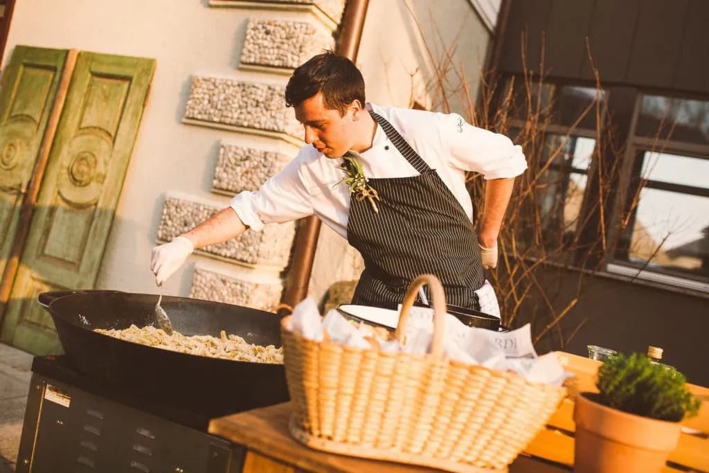 Chef stirring pasta in a large pan outdoors near a building with green doors, Dvor Jezersek.