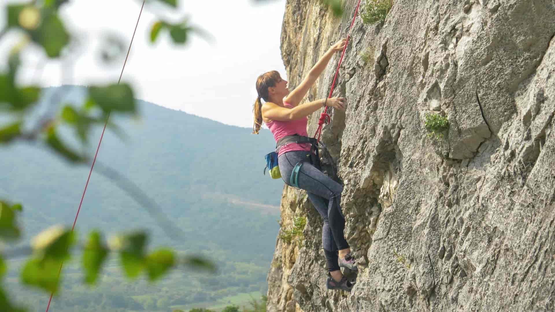 Woman rock climbing up a cliff face with green mountains in the Slovenian countryside.