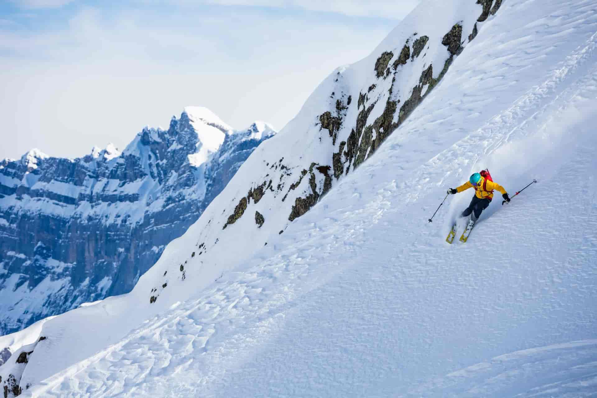 Skier descending steep snowy mountain slope with rugged snow-capped peaks in background