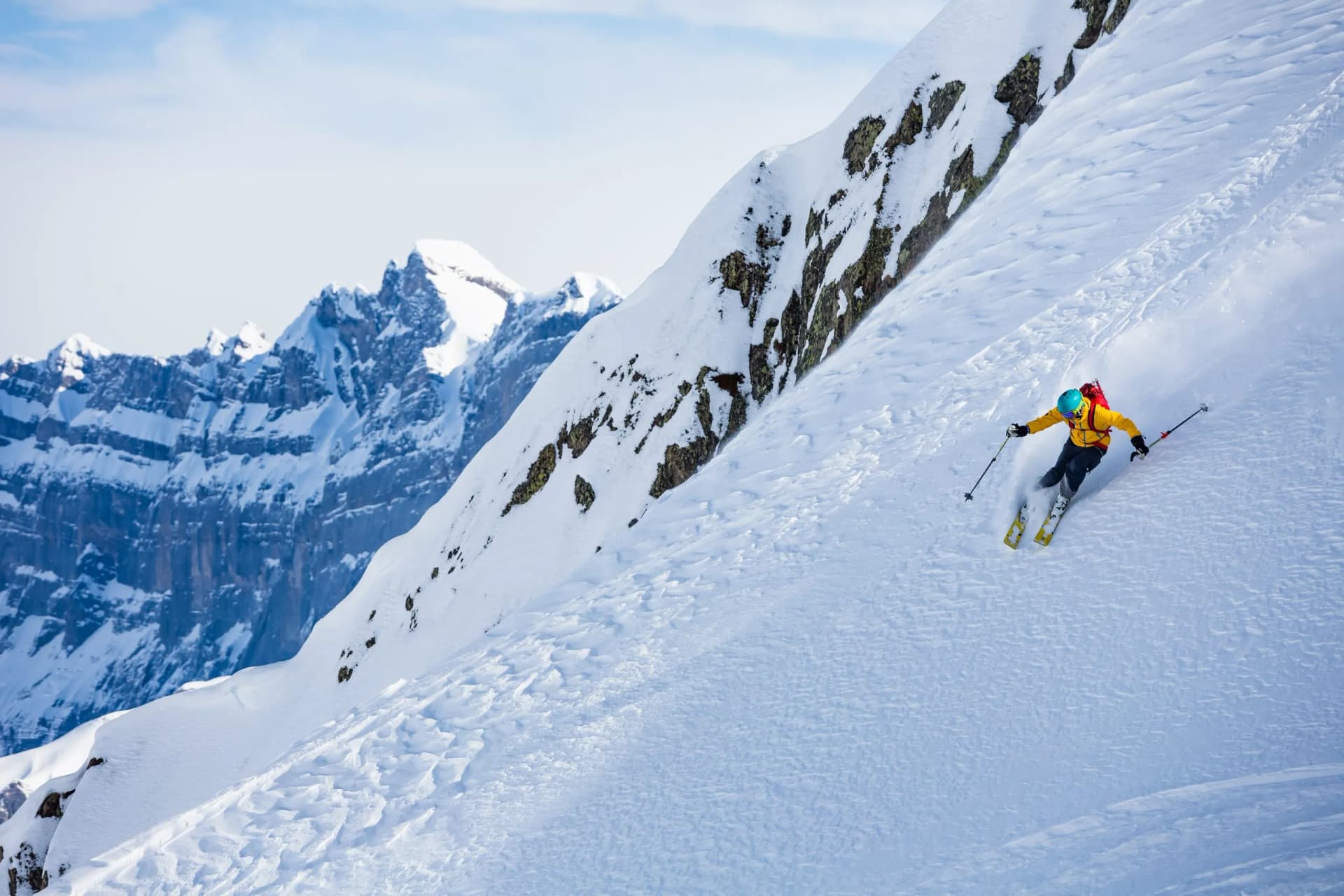 Skier descending steep snowy mountain slope with rugged snow-capped peaks in background
