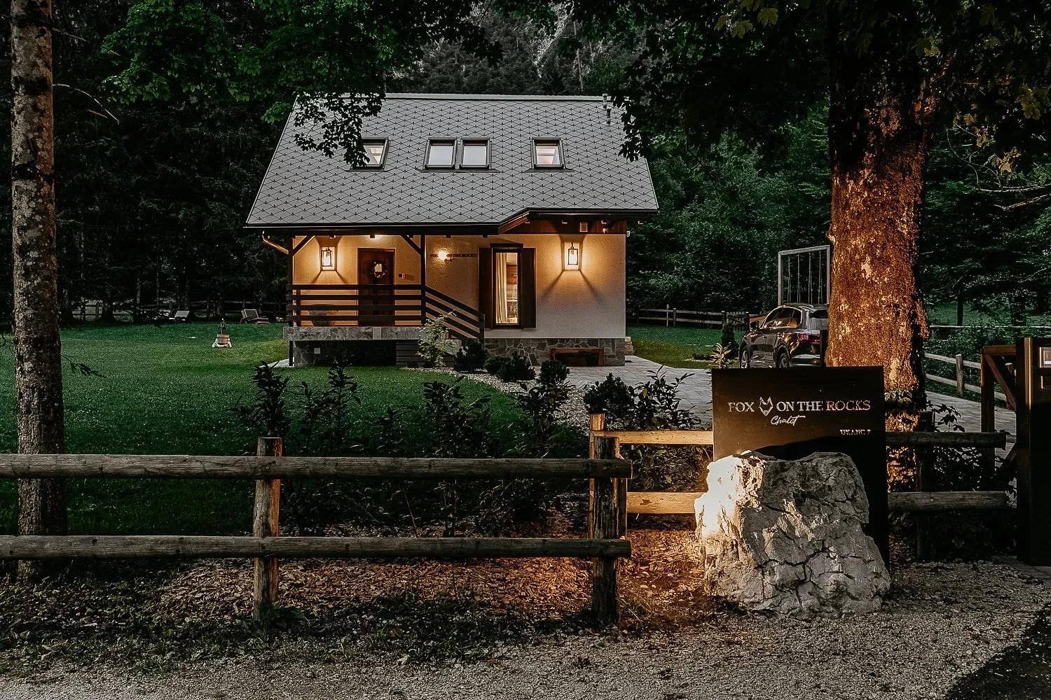 Chalet entrance at dusk with "Fox on the Rocks" sign, wooden fence, and surrounding forest.