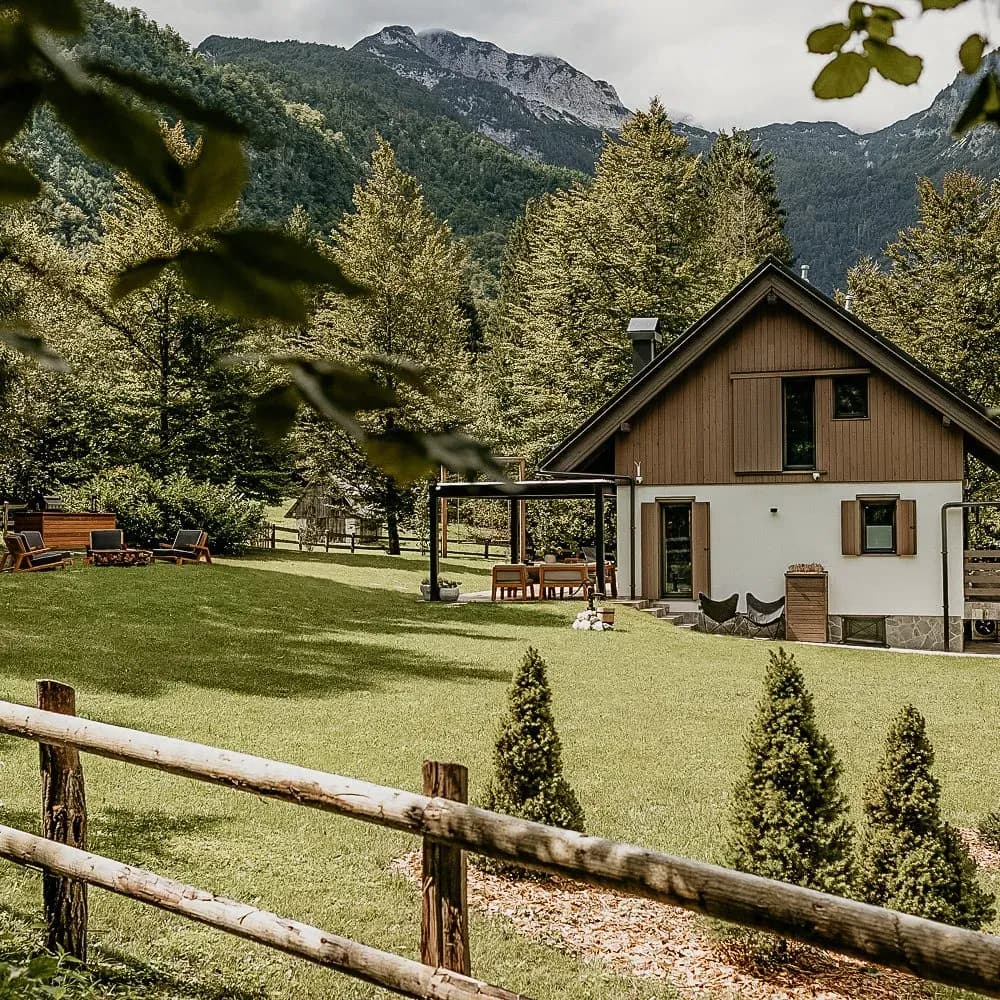 Chalet with white and wood siding in a grassy clearing, backed by forested mountains.