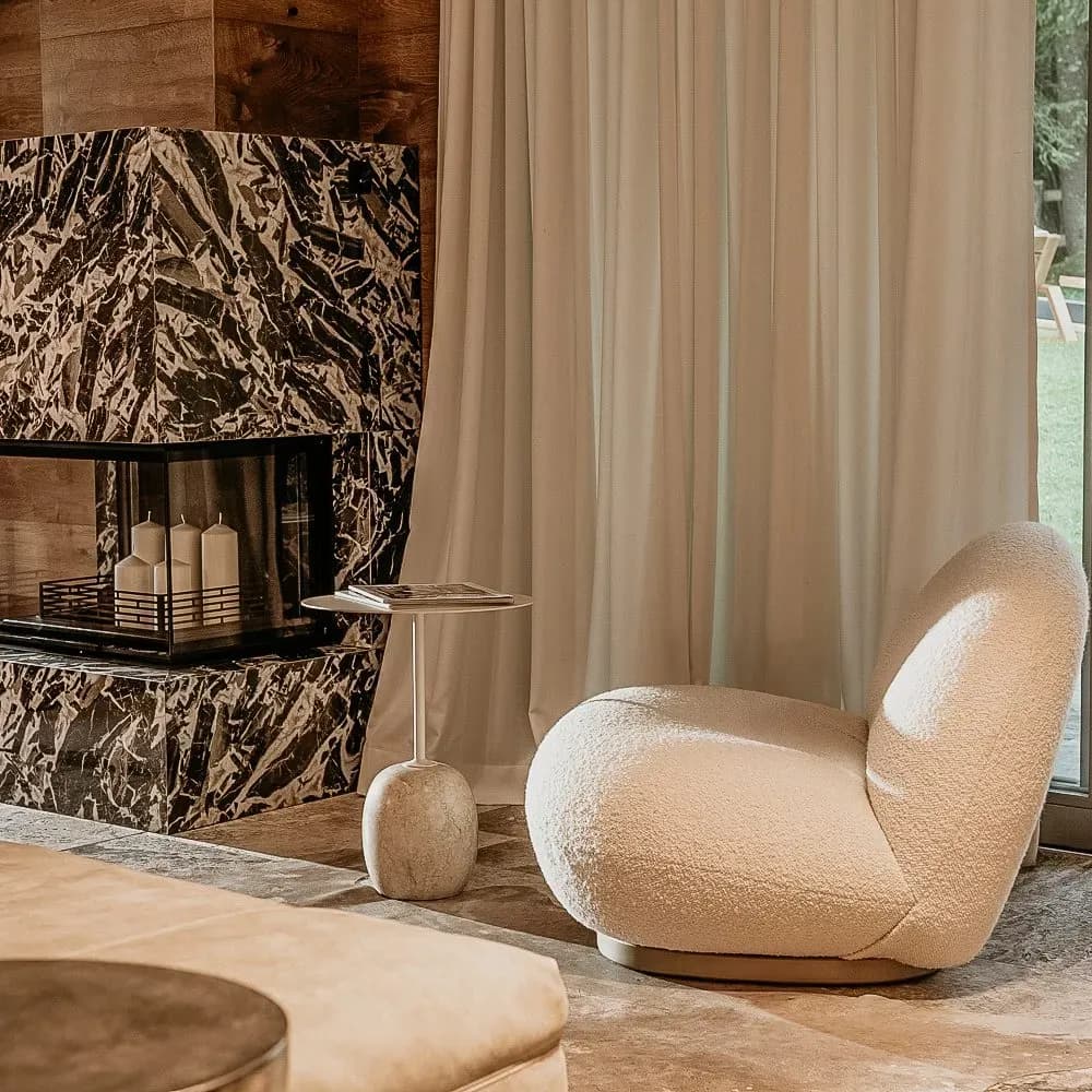 Cozy interior with a white boucle armchair next to a dark marble fireplace and light curtains.