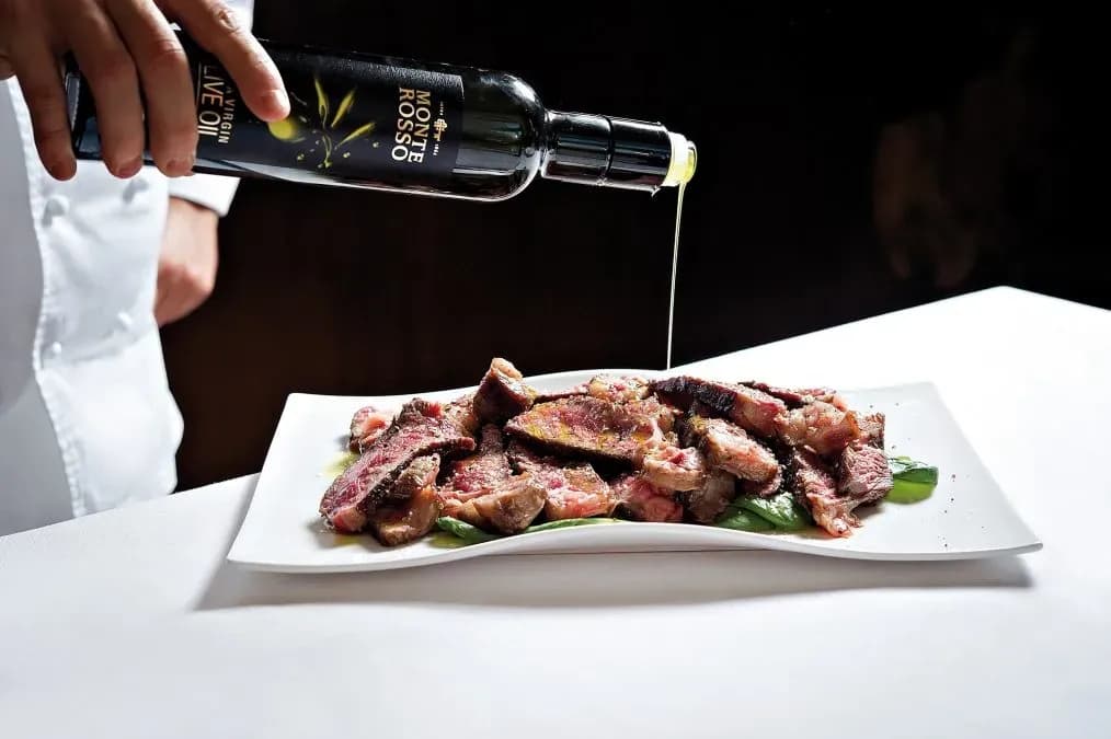 Chef pouring olive oil onto sliced steak served on greens on a white plate