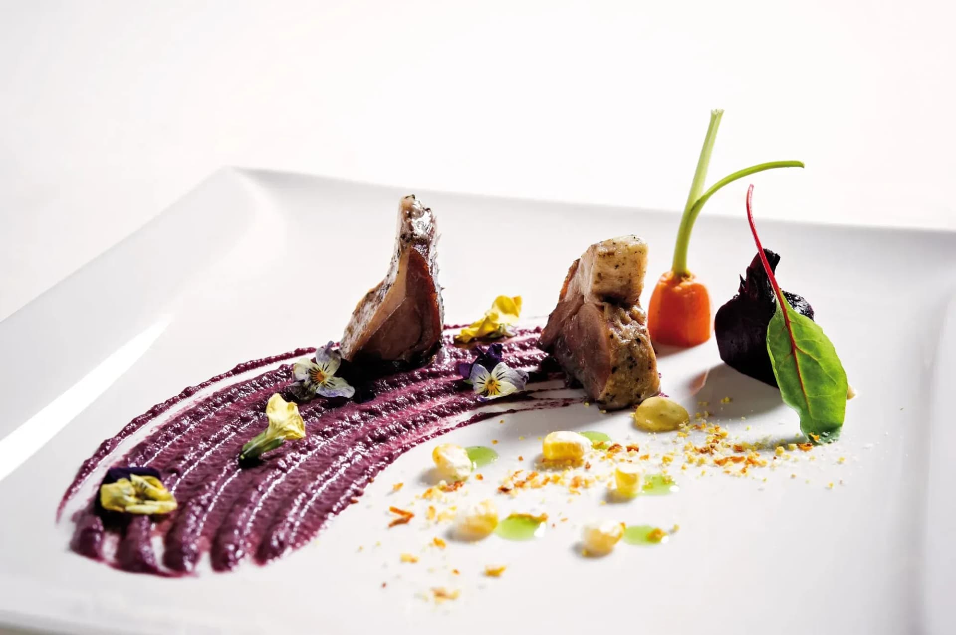 Gourmet dish with seared meat, purple puree, baby carrot, and edible flowers on white plate.