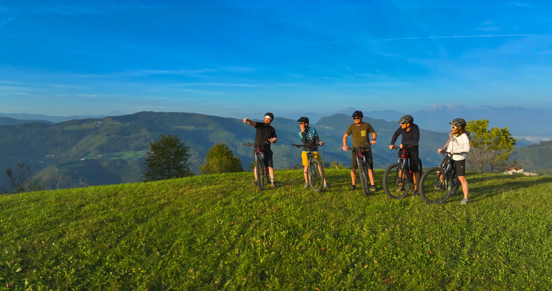 AERIAL: Cycling friends taking a break on a hilltop, surrounded by stunning panoramic mountain views. They are enjoying the beauty of the landscape after climbing uphill with their mountain bikes.