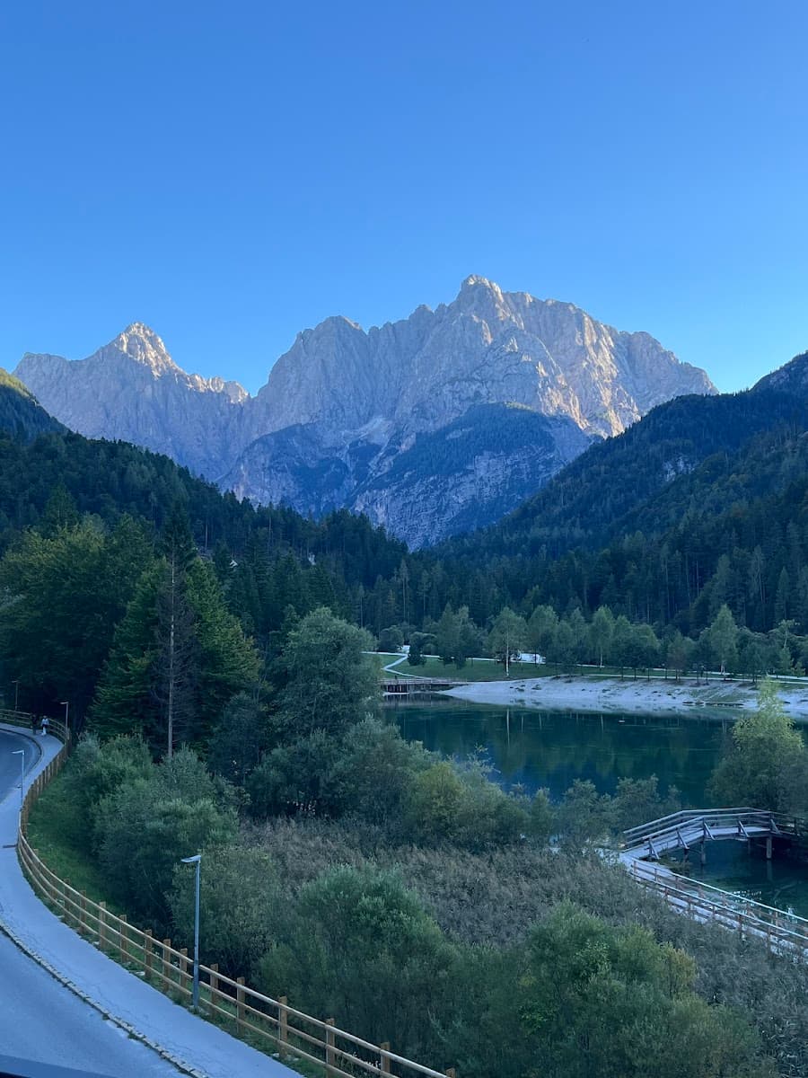 Jagged mountain peaks above a forest, lake, and paved path with wooden railing.