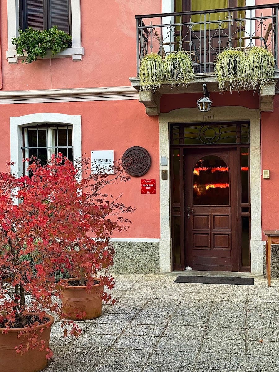 Entrance to a salmon-pink building with a dark wood door, potted red maple tree, and balcony.