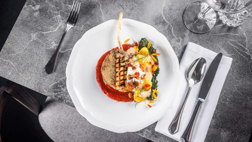 Grilled chop with bone on red sauce, vegetables, and cream on white plate at dining table