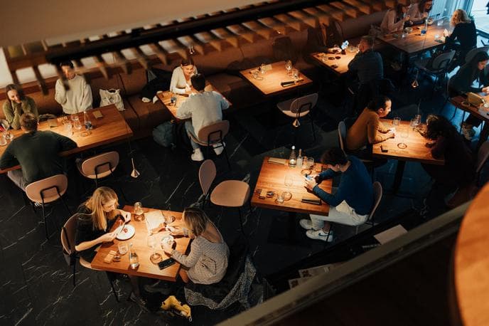 Diners seated at wooden tables inside a dimly lit, modern bistro or restaurant.