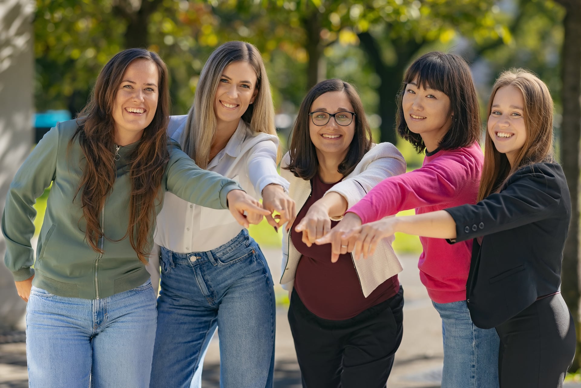 Five women smiling and pointing their fingers toward the camera in an outdoor park setting.
